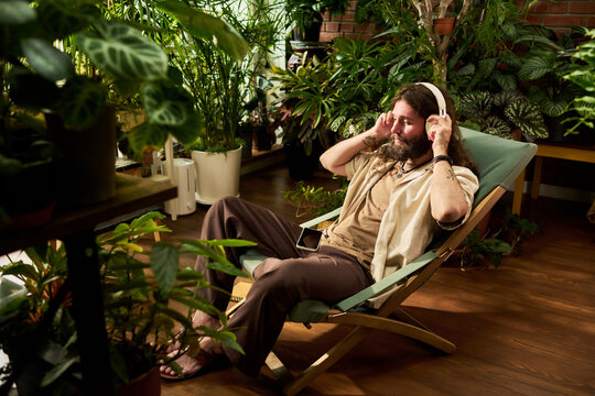 Young adult Caucasian man relaxing with wireless headphones in home greenhouse. Concept for mental wellbeing, mindfulness app advertising, music streaming service campaigns