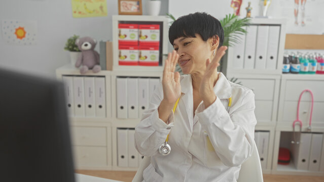 Woman doctor in white coat with yellow stethoscope raises fists and claps hands in a pediatric clinic building; telemedicine success joy.