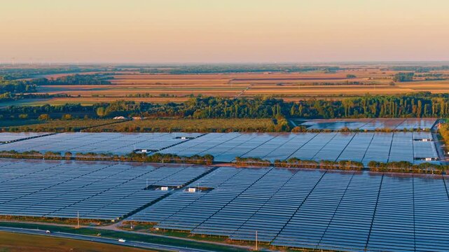 Solar panel rows, aerial view toward farmland at sunset. Photovoltaic arrays extend past service tracks and trees beneath a peach colored sky.