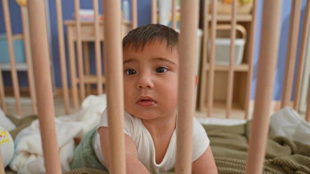 Toddler boy holding crib bars and reaching while sucking his finger on a blanket in a nursery inside a building; curiosity.