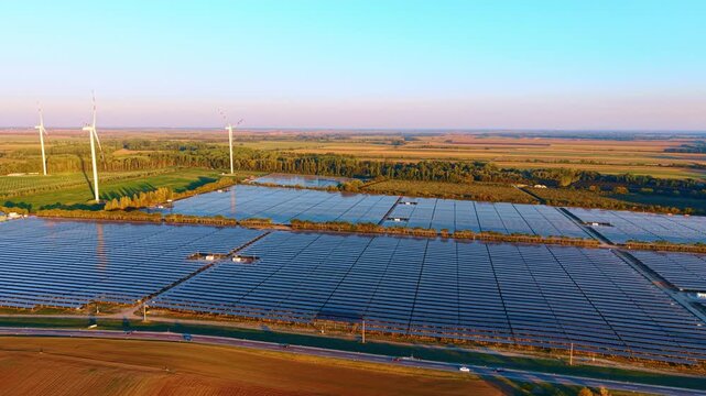 Solar farm and wind turbines, aerial view above a roadside installation. Panel blocks follow parallel lines, with brown fields below and a cyan sky above.