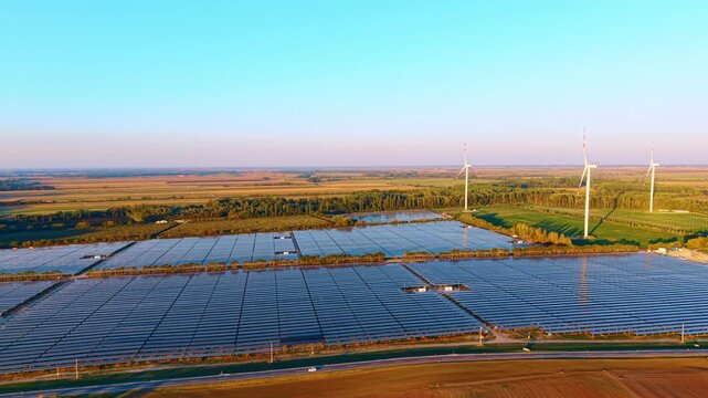 Solar energy farm surrounded by rural fields. Elevated view shows a large solar panel array with wind turbines standing in distant green fields.