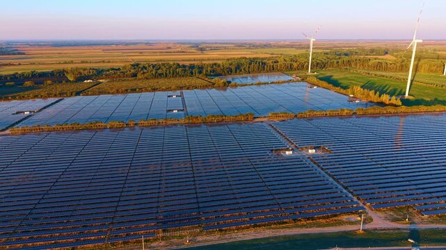 Solar farm and wind turbines, aerial view across aligned photovoltaic rows. Blue arrays surround service buildings, with tree belts and open fields on the horizon.