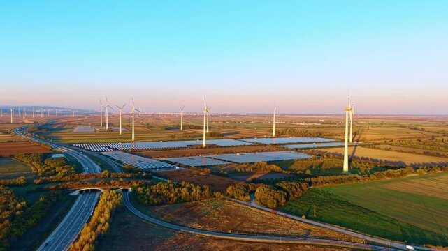 Solar arrays and wind farm, aerial panorama beside a highway. A curving road passes fields and tree belts below white towers under a clear blue sky.