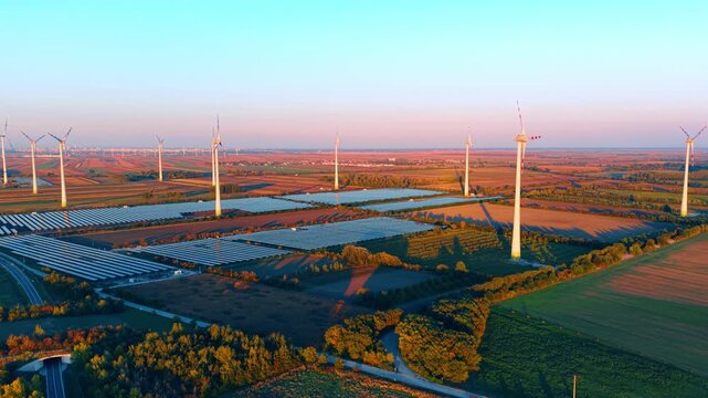 Solar farm and wind turbines, aerial view of energy sites among fields. White towers stand between blue arrays, with a road and dense trees in the foreground.