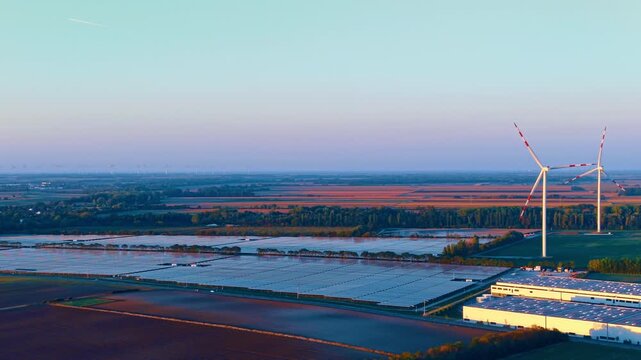 Solar panels and industrial building near wind turbines. Drone view shows solar panel fields beside a warehouse building with wind turbines under dusk sky.