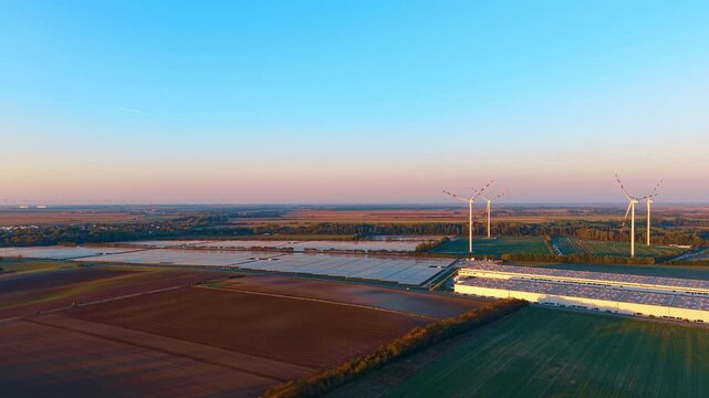 Solar farm and wind turbines, aerial view with industrial roofs. Brown and green fields fill the foreground beneath panel arrays and a broad cyan sky.