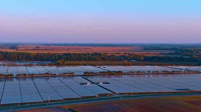 Solar panel farm in rural landscape at sunset. Aerial view shows blue photovoltaic arrays on green fields under purple sky.