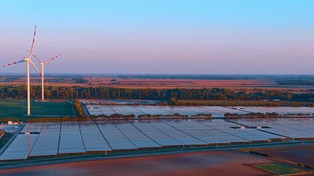 Solar arrays and two wind turbines, aerial view beneath an evening sky. Blue panel blocks extend beside green plots and trees, with red markings on the blades.