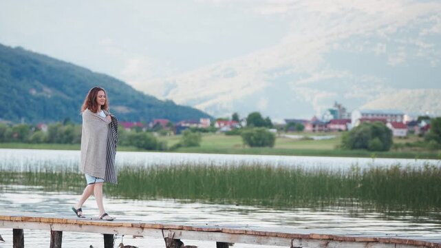 Young woman wrapped in a warm blanket walking along a wooden pier on a scenic mountain lake, enjoying peaceful nature and breathtaking mountain views.