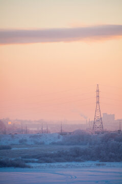 Winter energy background with a steel transmission tower and distant heating plant buildings behind a frosty valley under a pink hazy sunset glow