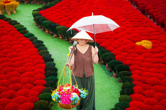 A woman in a conical hat with baskets among colorful incense sticks. Quang Phu Cau, Hanoi, Vietnam.
