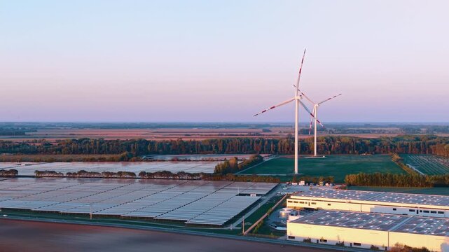Solar panels and two wind turbines, aerial view above an industrial site. A warehouse roof fills the lower right beside blue arrays and green plots beneath a pastel sky.