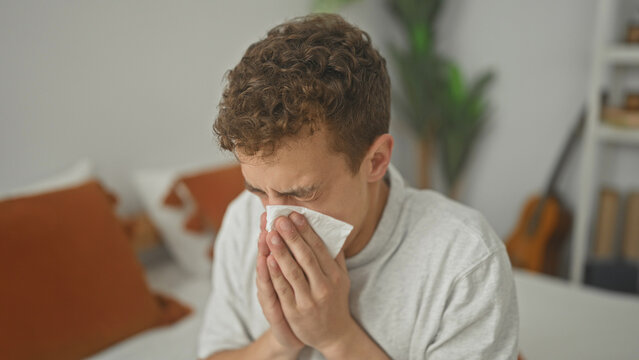 Young hispanic man on bed holding tissue to nose with both hands, eyes closed and sneezing in bright bedroom; discomfort recovery.