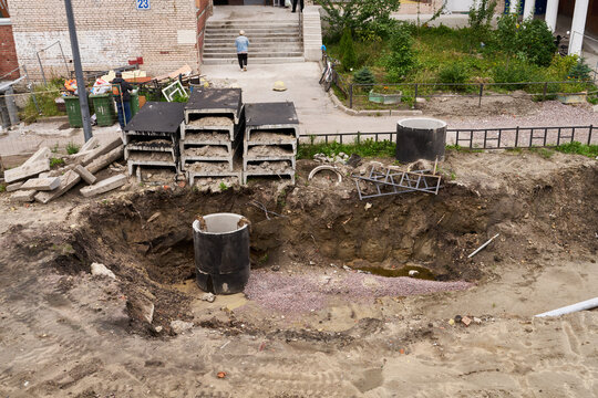 Open excavation at construction site with deep trench, large concrete rings, scattered pallets, and dirt around brick building backdrop, showing ongoing urban infrastructure work