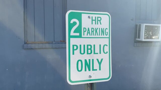 A green and white two hour parking sign mounted on a post against a blue painted brick wall