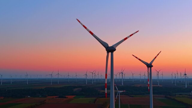 Wind turbines with red blade markings, aerial view at sunset. Two white towers rise above dark fields, with a pink horizon fading into a blue sky.