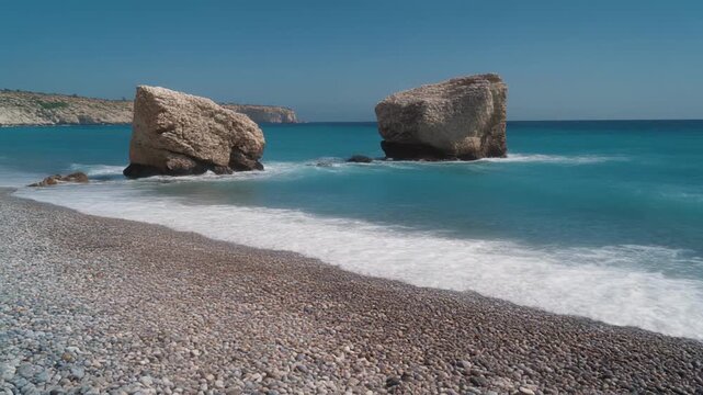 Ocean waves gently washing over a pebble beach with large rock formations under clear blue sky