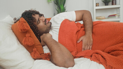 Young indian man clutching stomach and holding an orange knitted blanket while lying on bed with tassel pillow and white sheets inside building  discomfort recovery. © Krakenimages.com