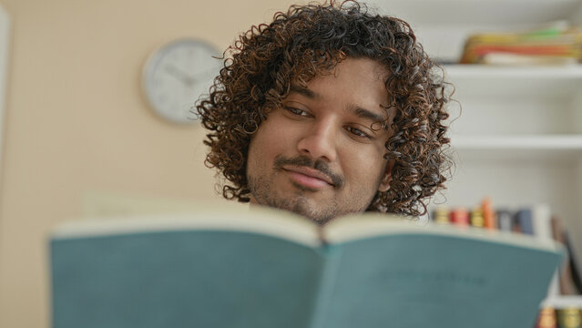 Young indian man reading teal book, hand on chin while turning page in a building with bookshelf and wall clock visible; contemplation.
