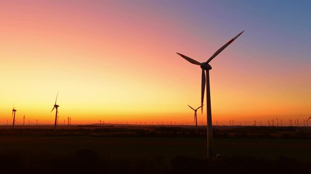 Wind turbine, backlit rotor above a rural wind farm at sunset. Dark towers punctuate the fields beneath a sky that grades from yellow to orange and blue.