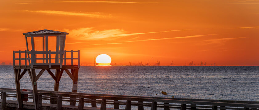 Luc-Sur-Mer, France - 08 08 2026:  Panoramic view of a wooden pier in front of the beach, sea, a colorful sunrise and Le Havre city far away