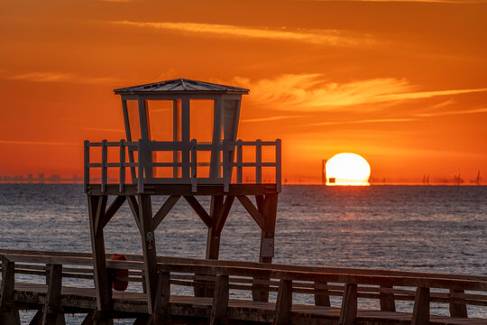 Luc-Sur-Mer, France - 08 08 2026:  Detail view of a wooden pier in front of the beach, sea, a colorful sunrise and Le Havre city far away