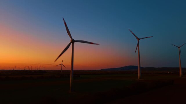 Wind farm operation during twilight blue hour. Spinning blades harvest natural kinetic power across dark fields as night falls.