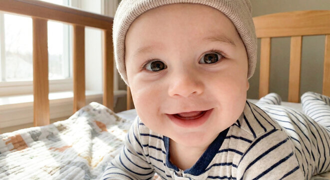 Close up of happy infant in striped pajamas smiling at camera while lying on soft blanket in bright cozy nursery.
