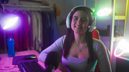 Young hispanic woman gamer wearing headset smiles, hand on desk near keyboard and microphone in a gaming studio  joyful streaming. © Krakenimages.com