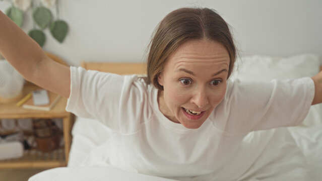 Woman sitting up in bed with arms outstretched and startled expression inside a bedroom building; surprise.