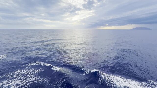 Ship bow waves with white foam on deep blue Mediterranean Sea under dramatic cloudy sky with golden light and island on the horizon