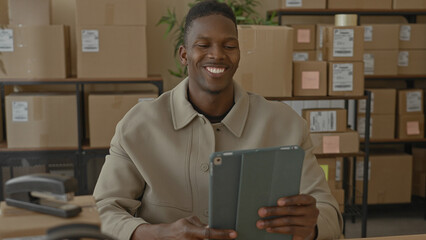 Man in tan jacket holding tablet waves hand and smiles beside stacked parcels on shelving in building  friendly greeting. © Krakenimages.com