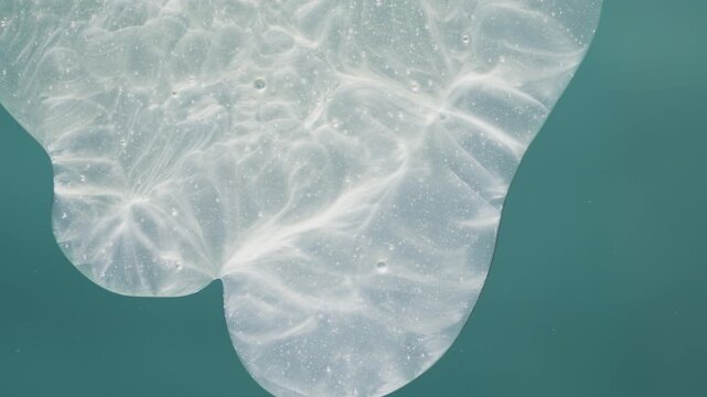 Shiny pearlescent cosmetic gel moving slowly. Sun glare and bubbles create a rippling texture on the transparent liquid on a green surface