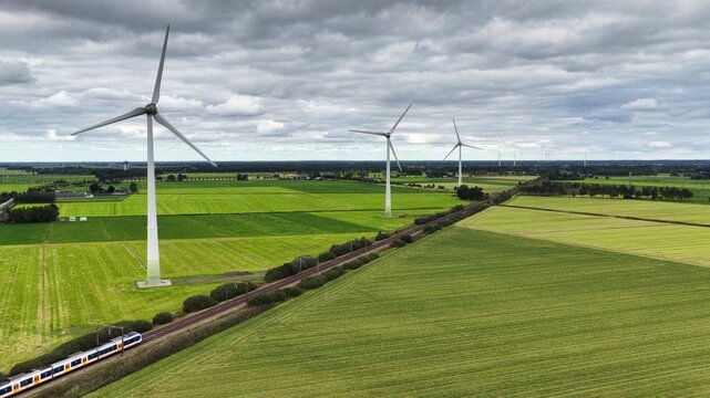 A passenger train of the Nederlandse Spoorwegen NS travels along wind turbines producing sustainable energy.