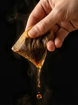 Tea bag, hand holding hot tea bag with dripping liquid and steam against dark background, morning beverage concept.