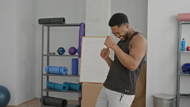 Man with bare arms shadowboxing and smiling, fists raised near yoga mats, foam rollers, dumbbells, boxing gloves and a whiteboard in a training studio; joy confidence focus.