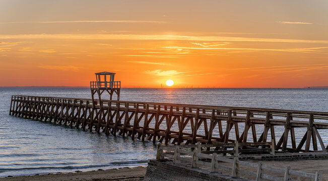 Luc-Sur-Mer, France - 08 08 2026:  Panoramic view of a wooden pier in front of the beach, sea, a colorful sunrise and Le Havre city far away