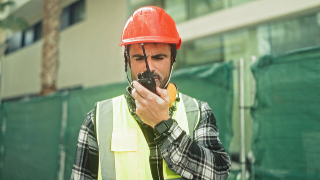 Man engineer in orange hardhat and reflective vest speaks into a walkie talkie and holds radio near building site; safety communication focus.