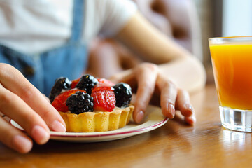 Female hands hold sweet cake with berries on table with juice in cafe - Hands hold berry tart on plate. Glass of orange juice sits nearby. Soft focus background enhances focus © Pavel Losevsky