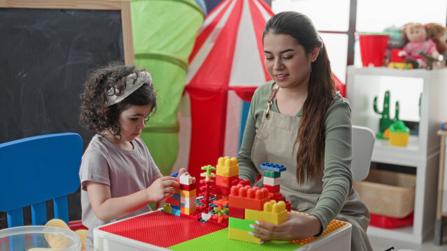 Woman teacher guides girl stacking colorful blocks with her hand on a play table inside a preschool building; nurturing learning.