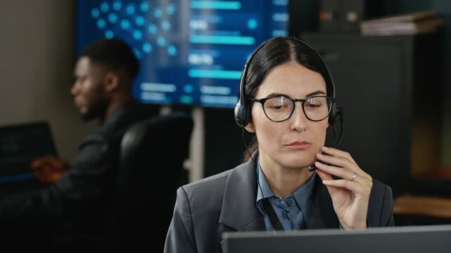 Young adult Caucasian woman wearing headset talking into microphone while working at computer in cybersecurity office. Clip fits tech support and network security promos