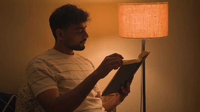 Young man sitting comfortably at home and reading a book at night under warm ambient lamp light. suitable for reading, education, learning, mindfulness, self-development and nighttime lifestyle.