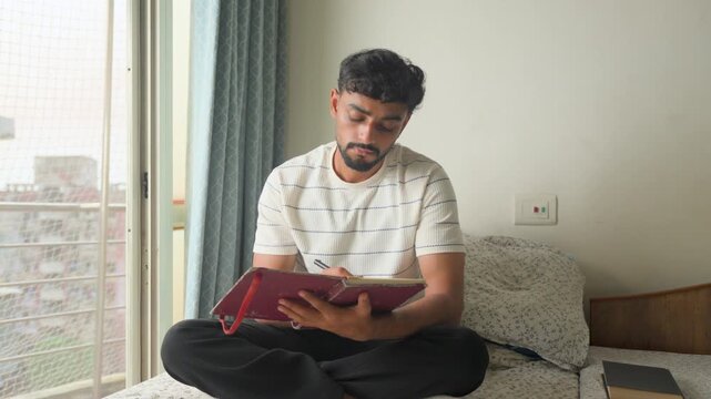 Young man sitting on a bed, holding a journal and thoughtfully reflecting while writing at home. suitable for journaling, self-reflection, personal development, mindfulness, creativity and planning.