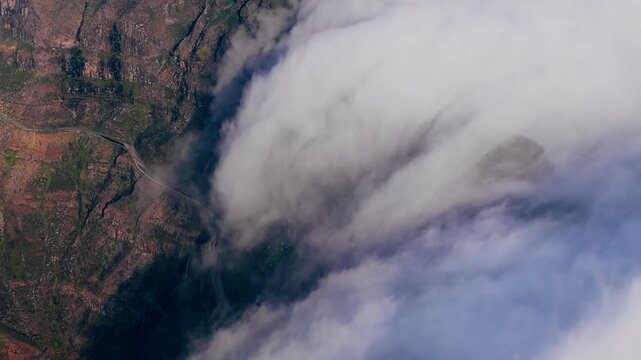 Encroaching clouds cascading down steep cliffs and engulfing mountain valley. Ominous aerial landscape with slopes disappearing beneath dense cloud cover