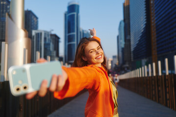 Naklejka na ściany i meble Smiling young woman in an orange jacket takes a selfie with a light blue smartphone, arm outstretched, against a city skyline of skyscrapers. Concept of mobile lifestyle and travel.