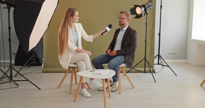 Journalist interviews guest with microphone in studio. Professional lighting and backdrop frame a seated conversation for news media and corporate content. Clear concept of media communication.