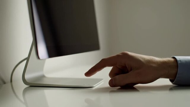 A person's hand reaches out to press the power button on the base of a sleek silver desktop monitor, turning the screen off in a minimalist office setting.