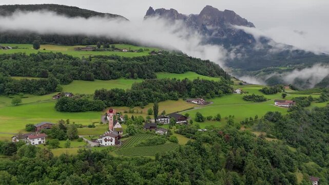 Alpine village below rugged mountain peaks and bands of low cloud in Dolomites, South Tyrol, Italy. Aerial view