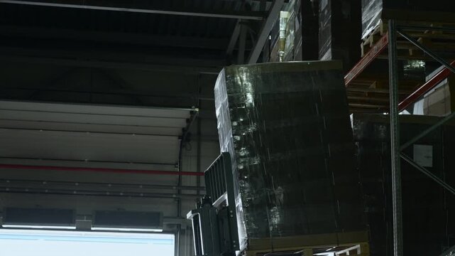 Dim industrial warehouse interior with tall metal storage rack, shrink wrapped pallet cargo, open loading doorway, and bright daylight creating strong contrast across dense logistics shelving